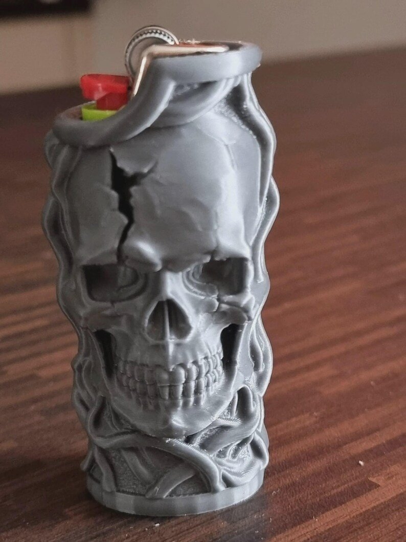 Skull & Roots Lighter Sleeve May include: A grey lighter case featuring a detailed, three-dimensional skull design with a prominent crack on the forehead. The skull is surrounded by swirling, vine-like textures. A red ignition button and a silver metal flint wheel are visible at the top.