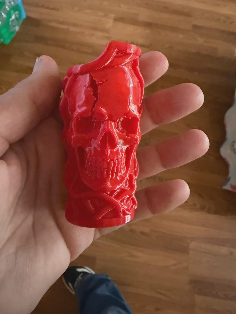 Skull & Roots Lighter Sleeve May include: A bright red, three-dimensional cylindrical object featuring a detailed skull design with prominent teeth, eye sockets, and a cracked texture on the forehead, surrounded by raised, vine-like patterns wrapping around the entire surface.