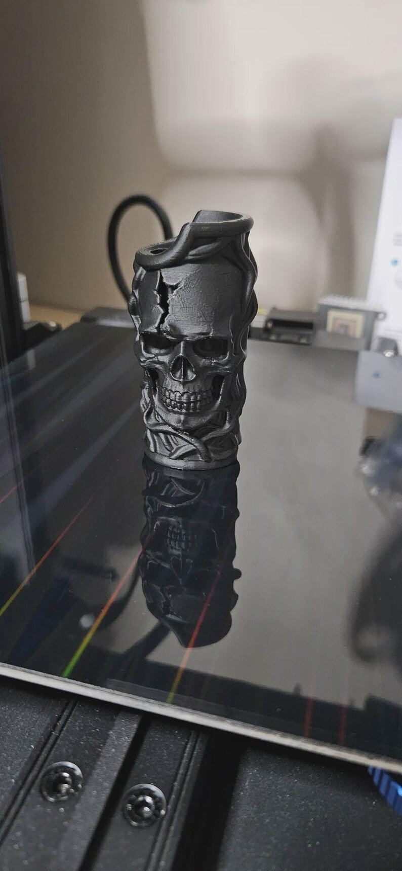 Skull & Roots Lighter Sleeve May include: A matte black 3D printed cylindrical object featuring a detailed human skull with a prominent crack on the forehead, surrounded by intertwined vine-like structures that wrap around the entire exterior surface of the vertical vessel.