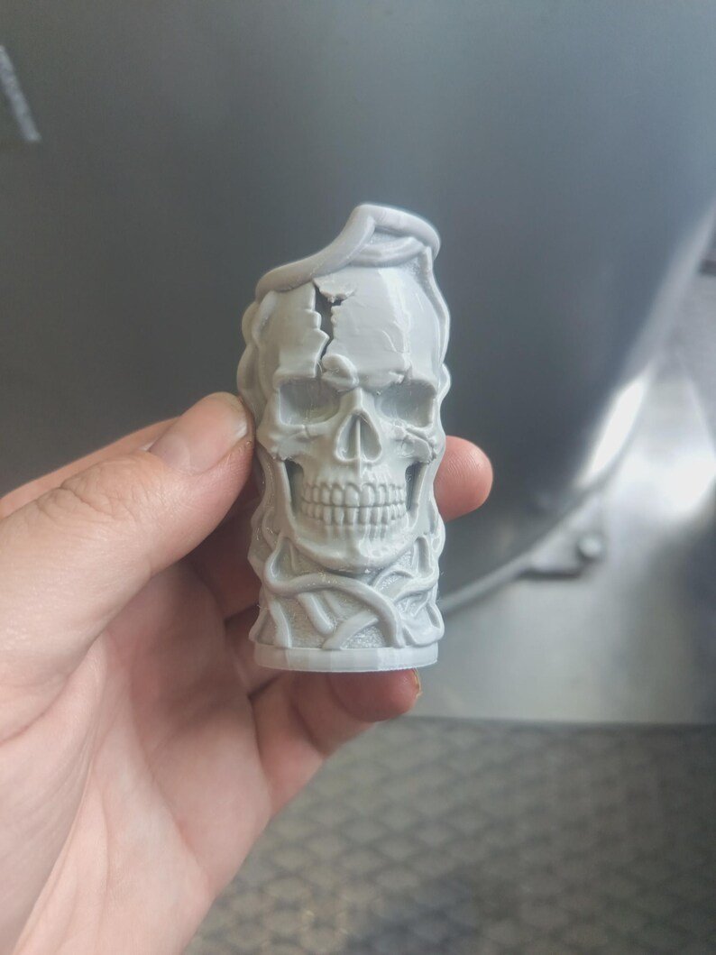 Skull & Roots Lighter Sleeve May include: A light grey, 3D-printed cylindrical object featuring a detailed human skull with a prominent crack on the forehead. The skull is surrounded by intertwined, vine-like structures that wrap around the base and top of the piece.