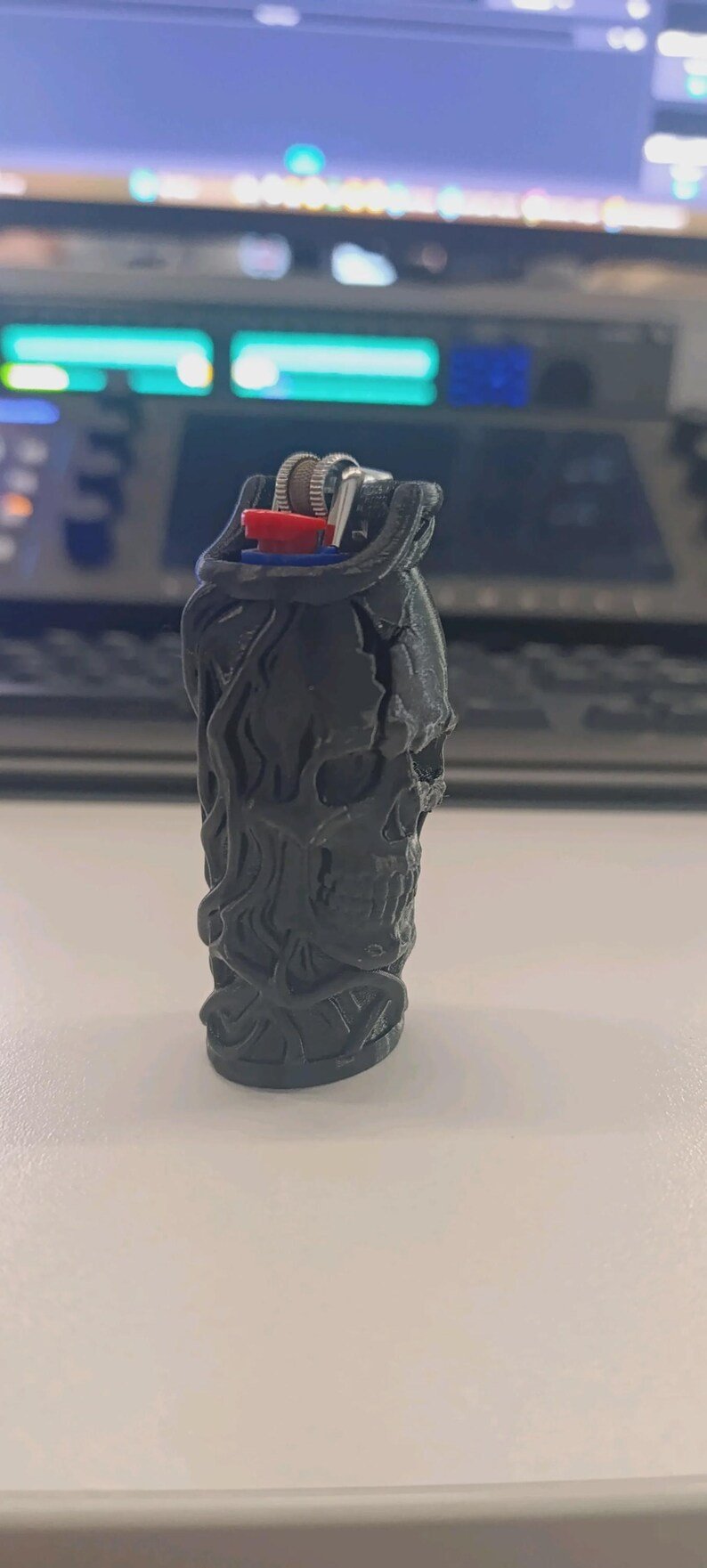 Skull & Roots Lighter Sleeve May include: A black lighter case featuring a three-dimensional skull design with intricate, flowing details. The case is moulded to fit a standard lighter, which has a visible red ignition button and a metallic silver flint wheel mechanism at the top.