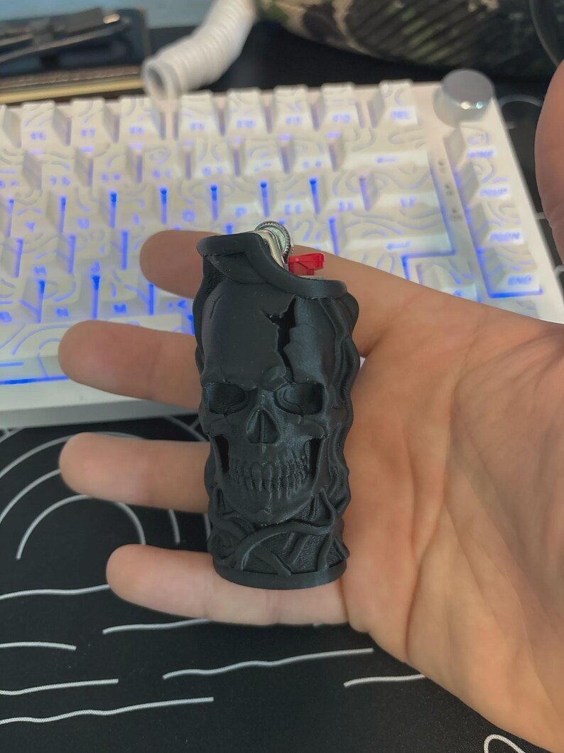 Skull & Roots Lighter Sleeve May include: A matte black lighter case featuring a detailed three-dimensional skull design with a cracked forehead and swirling vine-like textures surrounding the base. The case is designed to fit a standard rectangular lighter with a red ignition button.