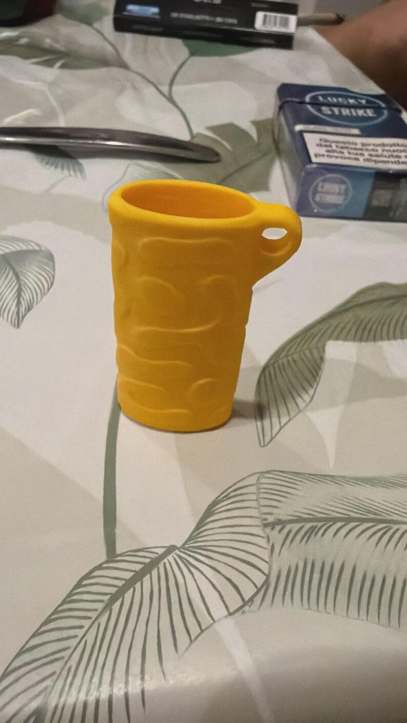 BIC Lighter Keychain Sleeve May include: A bright yellow, cylindrical silicone container featuring a textured, wavy surface pattern and a small, integrated side handle with a circular opening. The object is hollow and open at the top, designed with a uniform, matte finish.