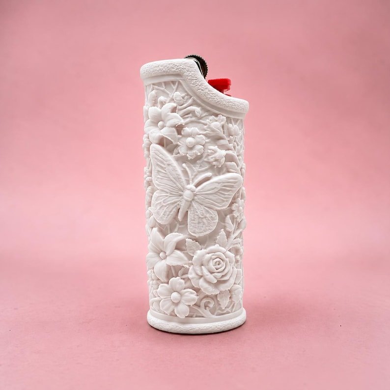Butterfly Garden Lighter Sleeve May include: A white lighter featuring a raised, textured relief design of a butterfly surrounded by various flowers and foliage. The top of the lighter shows a black ignition wheel and a small red plastic component visible at the top edge.