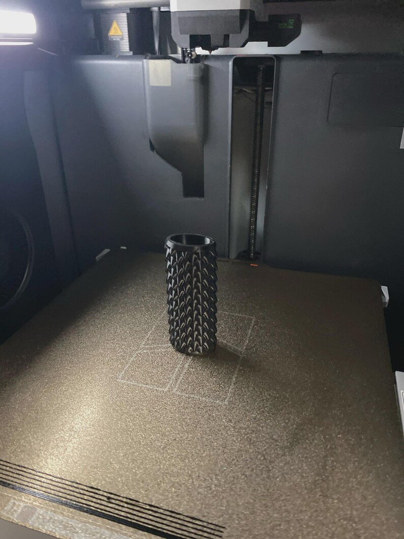 Smooth Dragon Scale BIC Lighter Sleeve May include: A black cylindrical object with a textured, spiked surface pattern standing upright on the metallic print bed of a 3D printer. The object features a repeating geometric design across its exterior, positioned centrally on the build plate.