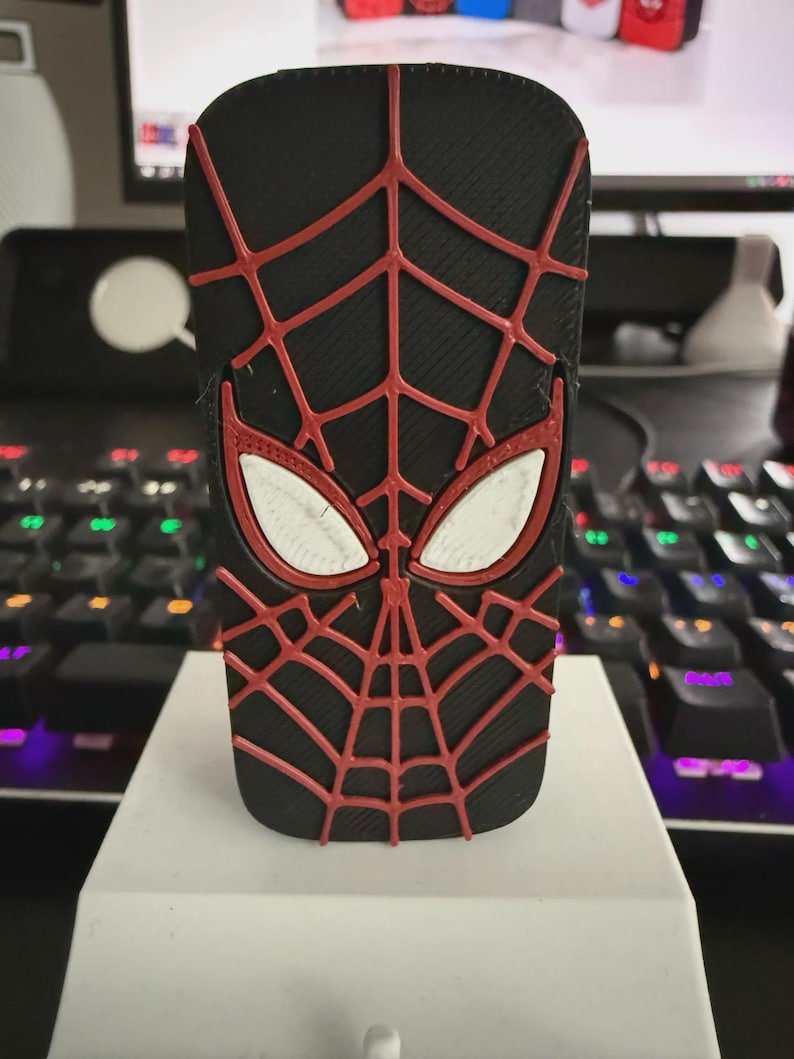 Spider-Inspired Lighter Case May include: A black rectangular object featuring a raised red web pattern and two white almond-shaped eye lenses. The design mimics a superhero mask with a textured surface, centred on a white base, showcasing a detailed three-dimensional graphic.
