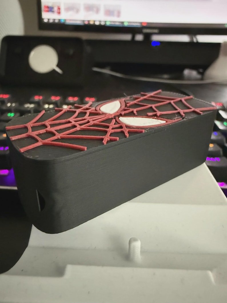 Spider-Inspired Lighter Case May include: A rectangular black plastic storage case featuring a raised red spiderweb pattern on the top lid. Two white, almond-shaped eye designs are integrated into the centre of the web pattern. The case has a smooth, matte finish.