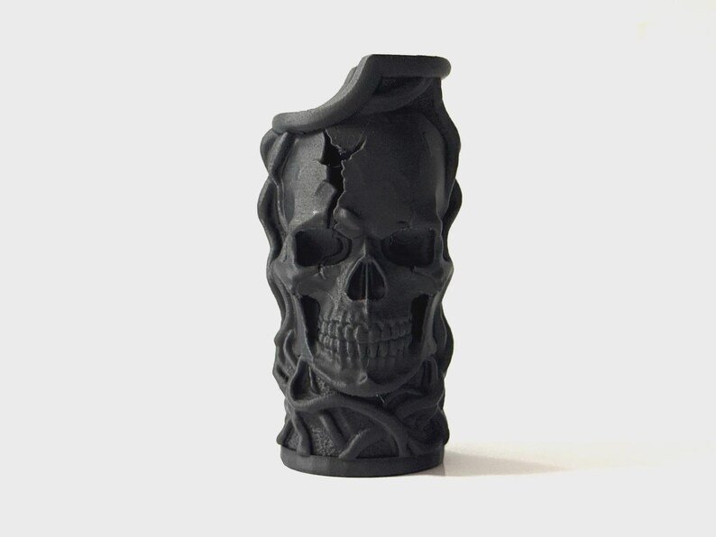 Skull & Roots Lighter Sleeve May include: A matte black cylindrical vessel featuring a sculpted human skull on the front surface. The skull design includes a prominent crack across the forehead and is surrounded by intricate, raised vine-like or root-like textures.