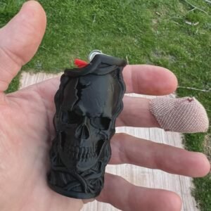 May include: A black lighter case featuring a three-dimensional moulded skull design with intricate bone detailing. The matte black accessory is shaped to fit around a standard lighter, with a red ignition button visible at the top of the casing.