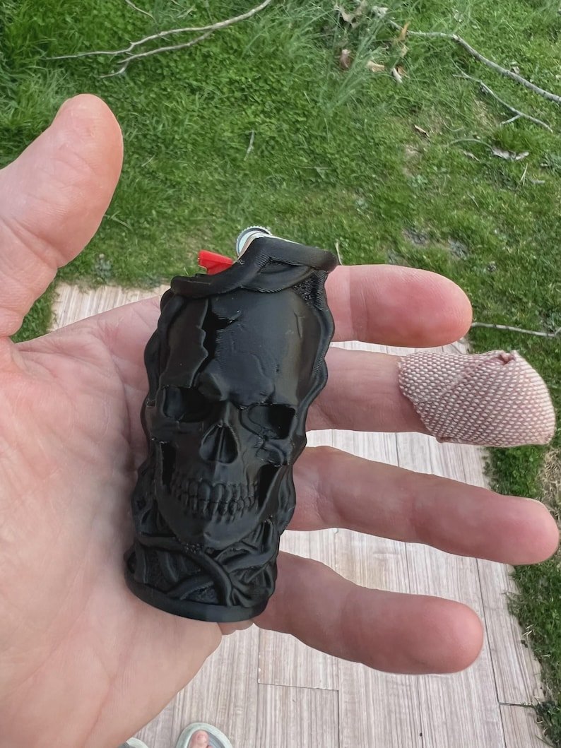 Skull & Roots Lighter Sleeve May include: A black lighter case featuring a three-dimensional moulded skull design with intricate bone detailing. The matte black accessory is shaped to fit around a standard lighter, with a red ignition button visible at the top of the casing.