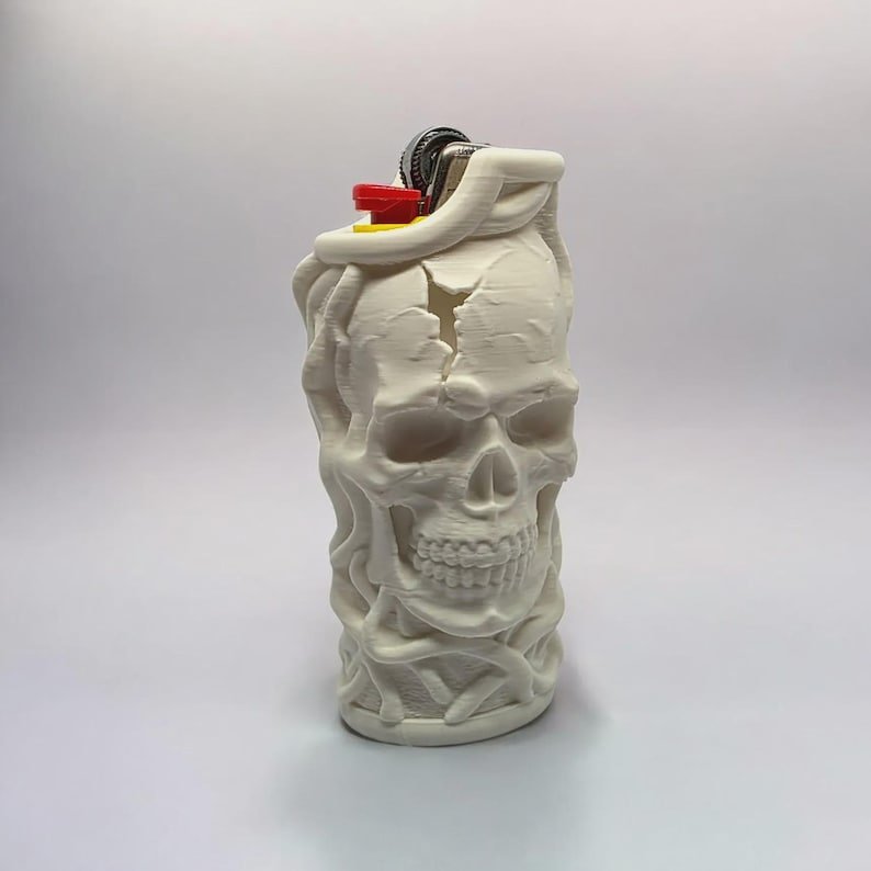 Skull & Roots Lighter Sleeve May include: A white 3D-printed lighter case featuring a detailed skull design with cracks on the forehead and textured vine-like patterns surrounding the base. The case holds a standard lighter with a visible red ignition button and metal top.
