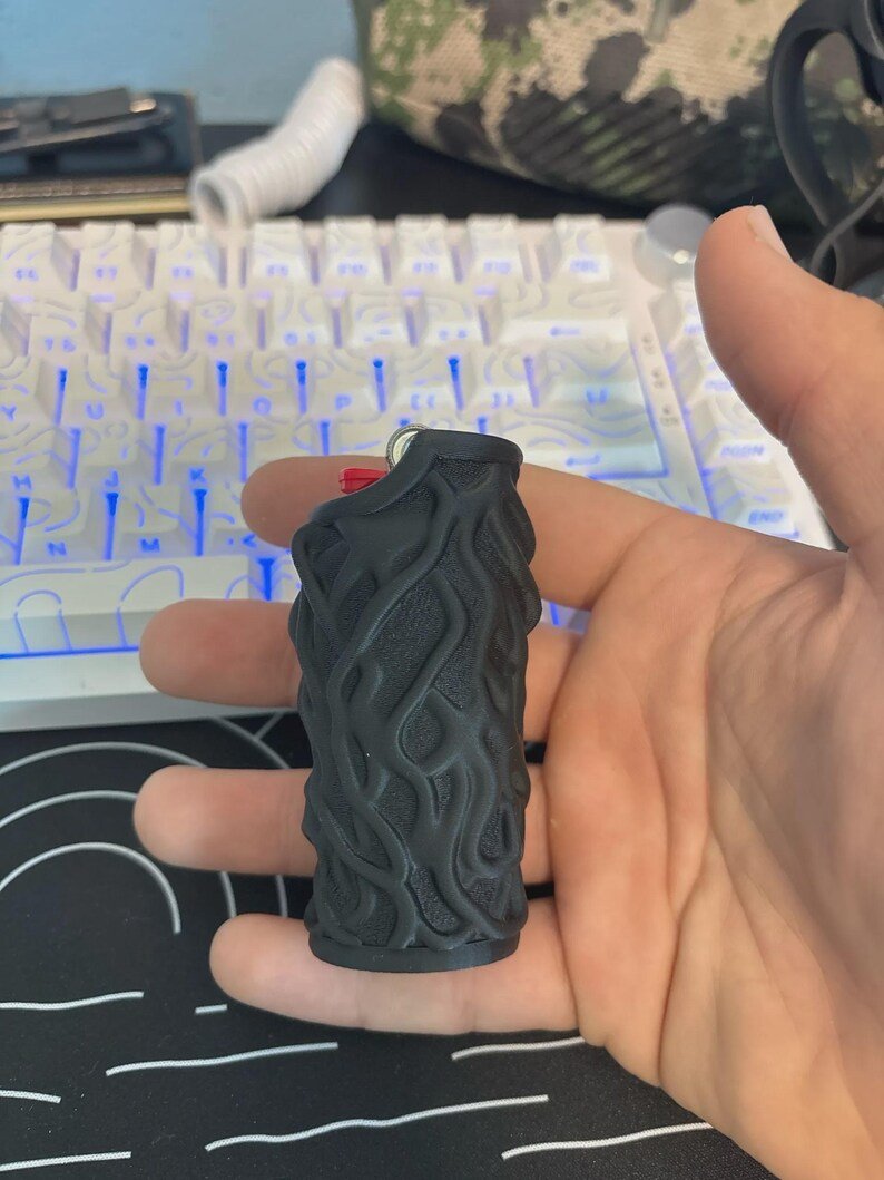 Skull & Roots Lighter Sleeve May include: A black lighter case featuring a raised, textured, organic vine-like pattern across its surface. The cylindrical sleeve is designed to fit over a standard lighter, with a small cutout at the top revealing the red ignition button.