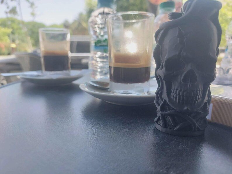 Skull & Roots Lighter Sleeve May include: A matte black, three-dimensional skull figurine with intricate, vine-like textures wrapping around its surface. The skull is positioned in the foreground, while two small glass cups filled with dark coffee sit on white saucers behind it.