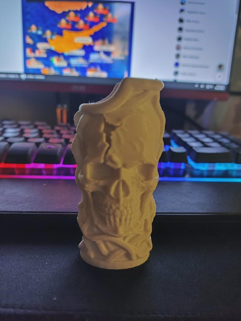 Skull & Roots Lighter Sleeve May include: A cream-coloured, 3D-printed cylindrical object featuring a detailed skull design integrated with swirling, vine-like patterns. The surface has a matte texture with visible layer lines, and the top edge is unevenly shaped.