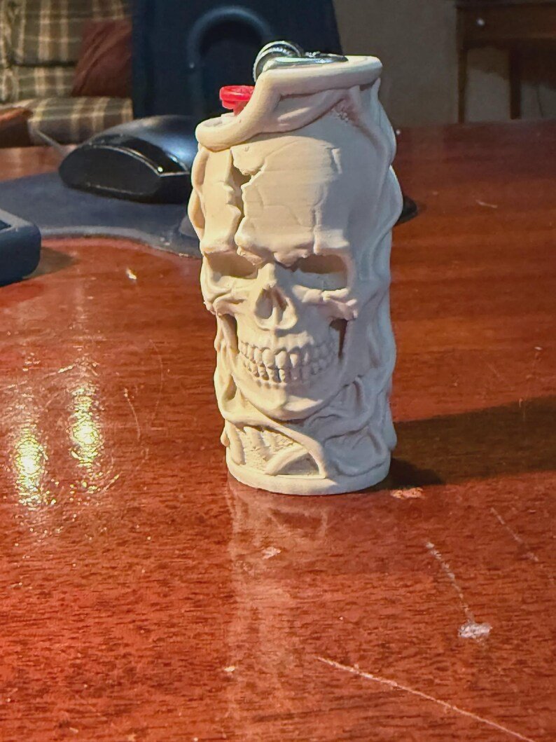 Skull & Roots Lighter Sleeve May include: A cylindrical lighter case featuring a detailed, off-white, sculpted skull design with cracked textures and swirling patterns. The top of the case reveals a small red ignition button and a metallic silver lighter mechanism.